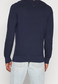 Homme portant un pull en maille bleu foncé et un jean bleu clair, vu de dos sur un fond blanc uni.
