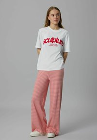 Vit T-shirt med fet röd text som säger "skulptur" och "KOLLEKTIV KLÄDER UTGÅVA", i kombination med röda och vita randiga vida byxor.