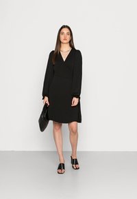 Vestido preto envolvente com mangas bufantes longas, tecido texturizado, decote em V e comprimento até a metade da coxa, combinado com sandálias pretas de slide e uma bolsa acolchoada.