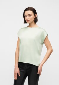 Νεαρή γυναίκα με κοντά σκούρα μαλλιά, φορώντας ανοιχτό πράσινο κοντομάνικο μπλουζάκι και μαύρο δερμάτινο παντελόνι, στέκεται μπροστά σε ένα απλό λευκό φόντο.