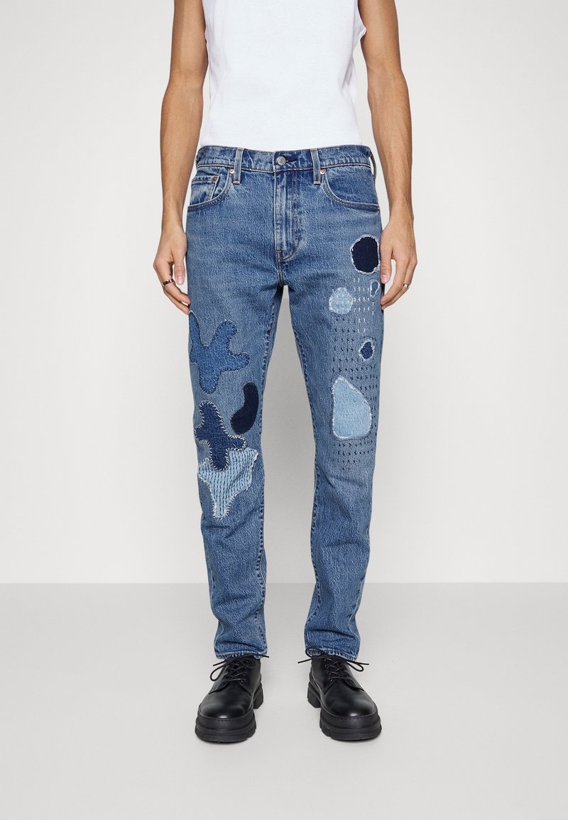 Homme portant un jean denim bleu orné de divers patchs cousus de teintes bleu foncé et clair, associé à des chaussures noires à lacets et un haut blanc sans manches.