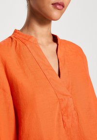 Femme portant un chemisier en lin orange vif à col en V avec un col bande, montrée des épaules aux lèvres sur un fond neutre.