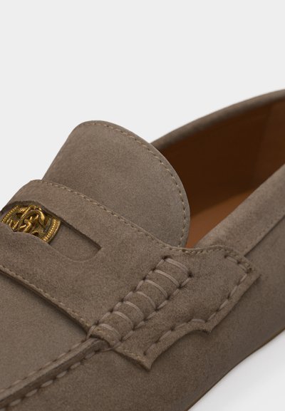 Mocassin en daim taupe avec un emblème décoratif doré sur la bride et des coutures détaillées le long des côtés et du dessus.