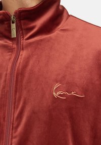 Vinröd zip-up-jacka av mjukt tyg med en guldembryderad logotyp på bröstet och hög krage.