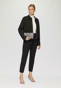 Zwarte jas, witte coltrui, zwarte broek, zilveren sandalen met hakken. Houdt een gestructureerde zilveren clutch vast. Minimalistisch ontwerp, moderne uitstraling.