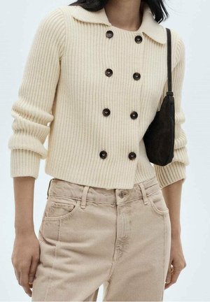Kremowy prążkowany kardigan dzianinowy z sześcioma ciemnymi guzikami, noszony z jasnobeżowymi spodniami i małą czarną torebką na ramię.