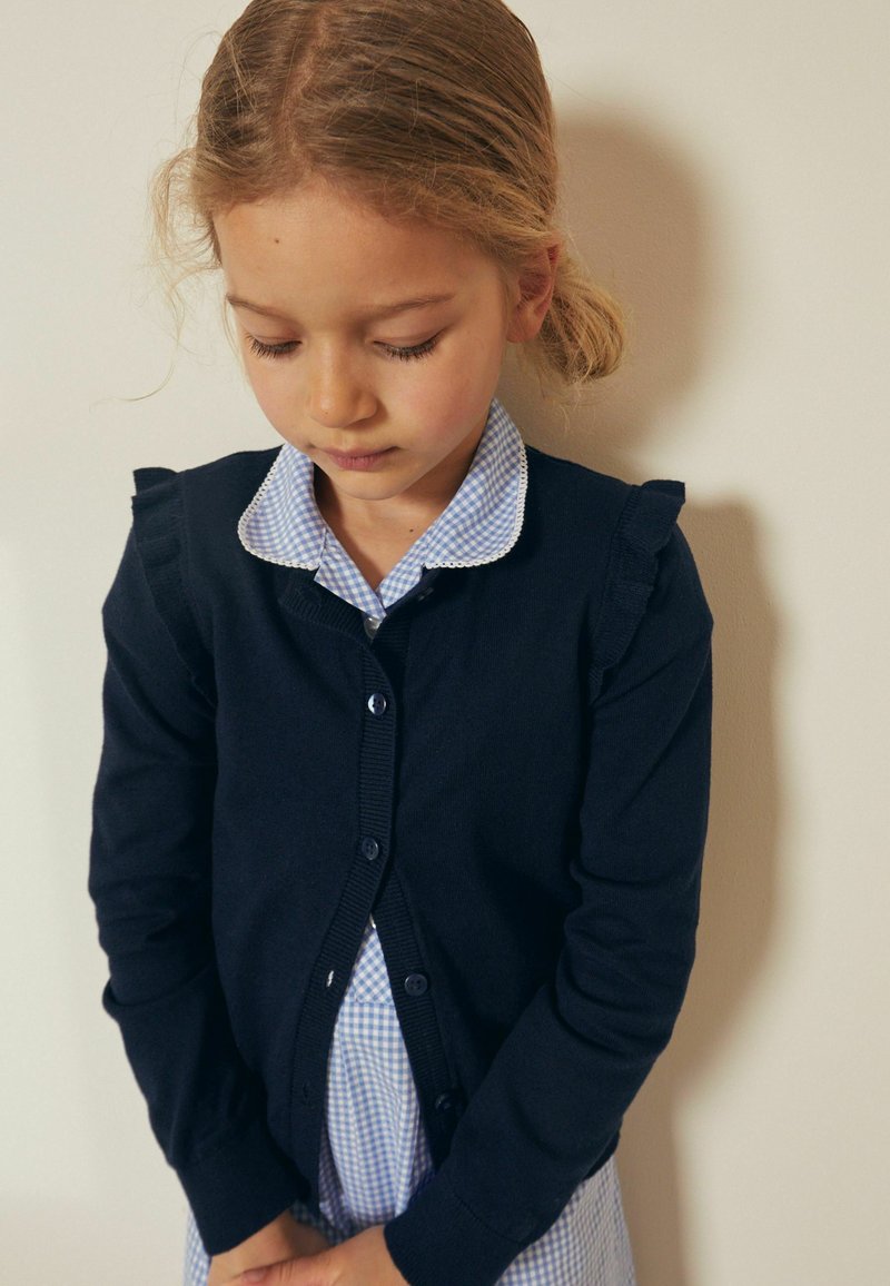 Navy cardigan with ruffled shoulders, button closure, and long sleeves, worn over a blue and white gingham shirt. Smooth texture.