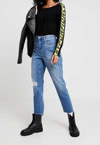 Top nero in rete a maniche lunghe con dettagli grafici giallo neon, abbinato a jeans blu strappati a gamba dritta e stivali da combattimento neri con lacci.