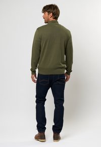 Suéter verde oliva con cuello alto, textura acanalada y mangas ajustadas, combinado con jeans de mezclilla oscura y botas marrones.