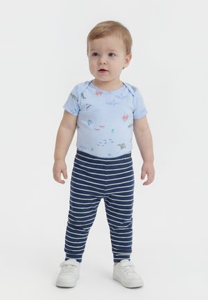 Μικρό παιδί που στέκεται, φορώντας ανοιχτό μπλε μπλουζάκι με κοντά μανίκια και σχέδια θαλάσσιων ζώων, μπλε σκούρο ριγέ παντελόνι και λευκά αθλητικά παπούτσια.