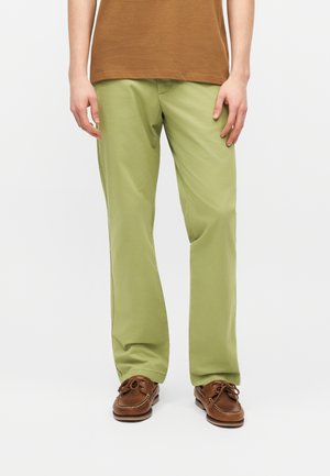 Person iført lysegrønne bukser med lige ben, brune bådsko og en brun kortærmet skjorte, stående mod en hvid baggrund.