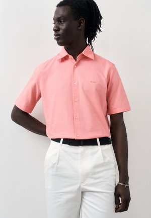 Mężczyzna o ciemnej karnacji i dredach, ubrany w różową krótko rękawową koszulę zapinaną na guziki oraz białe spodnie z czarnym plecionym paskiem.