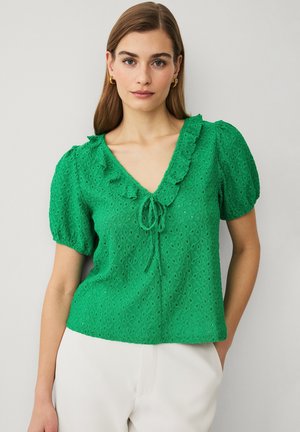 Frau trägt eine grüne, strukturierte Bluse mit Rüschenv-Ausschnitt und Bindendetail, kombiniert mit weißen Hosen, steht vor einem schlichten hellen Hintergrund.