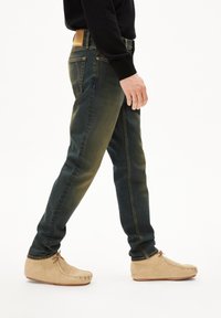 Donkerblauwe jeans met een vervaagde textuur en rechte pasvorm, gecombineerd met beige suède moccasinachtige schoenen.