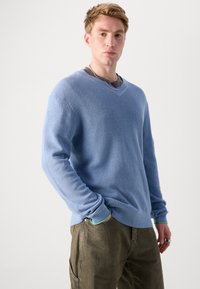 Jasnoniebieski sweter w serek z teksturowanego splotu, z długimi rękawami i ściągaczami. Noszony na szarym t-shircie, zestawiony z oliwkowymi spodniami.