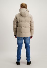 Beige pufferjack met een capuchon, met een gequilt ontwerp en geribde manchetten, gedragen over blauwe jeans en zwarte schoenen.