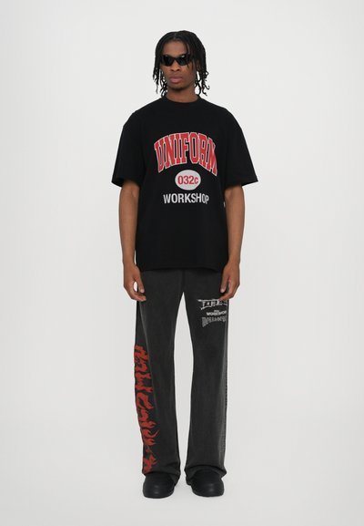 Camiseta negra de gran tamaño con gráficos en rojo y blanco en negrita, combinada con pantalones oscuros grises que presentan un patrón de llamas rojas y detalles impresos. Calzado negro.