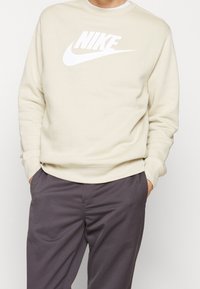 Beige sweatshirt med en vit Nike-logotyp, rundad halsringning, ribbad mudd och nederkant; parat med mörkgrå byxor. Slät textur.