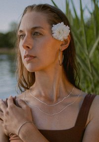 Silver necklaces layered on a person wearing a brown top. A large white flower is tucked behind their ear; they have a silver bracelet on their wrist.