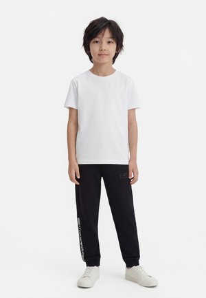 Kind staat en draagt een effen wit T-shirt met korte mouwen, zwarte joggers met zijlogo en witte sneakers op een witte achtergrond.