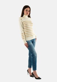 Maglione a collo alto crema con righe orizzontali beige, dotato di bottoni decorativi sulle spalle; abbinato a jeans blu aderenti e tacchi neri.