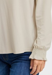 Beige langærmet skjorte med afslappet pasform. Har smocked manchetter og en glat tekstur, parret med mørkeblå denimjeans.