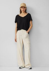 T-shirt noir oversize à col en V, pantalon cargo large crème avec poches latérales, et casquette beige ; chaussures de sport argentées.