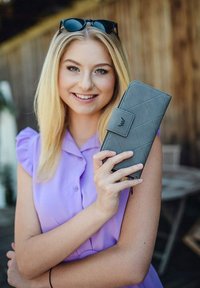 Gray quilted wallet with a clasp, held in a hand. Flat rectangular shape, featuring a logo and textured surface. Purple blouse in the background.