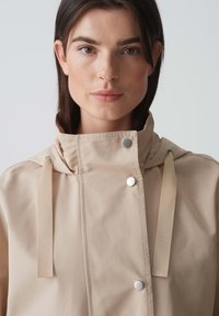 Giacca impermeabile beige con colletto alto, bottoni a pressione e cinturini regolabili. Texture liscia con un design minimalista.