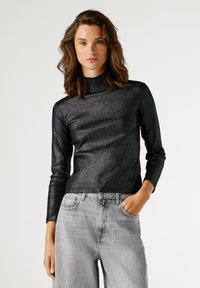 Νεαρή γυναίκα με κυματιστά καστανά μαλλιά, φορώντας μαύρο μεταλλικό τύπου ζιβάγκο και ανοιχτό γκρίζες ψηλόμεσες τζιν, ποζάροντας με το ένα χέρι στη τσέπη.