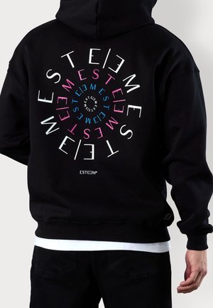 Person iført sort hættetrøje med flerfarvet spiraltekst "ESTEEM" på ryggen i hvide, lyserøde og blå bogstaver, lagdelt over sorte bukser.