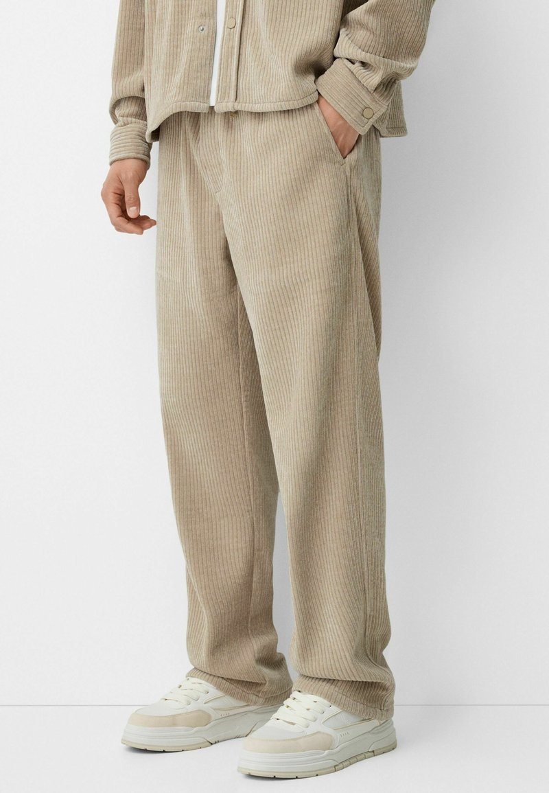 Pantalones de pana beige con un corte relajado y rayas verticales, que cuentan con bolsillos laterales y una cinturilla sin costuras, combinados con zapatillas blancas.