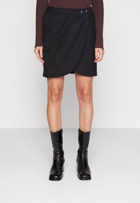 Svart wrapkjol med en slät textur, två framfickor och dekorativ hårdvara i midjan. Bärs med svarta ankelskor.