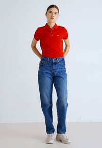 Camisa polo roja con cuello, combinada con jeans de mezclilla azul de tiro alto. Modelo que lleva zapatillas de color claro, en un fondo neutro.