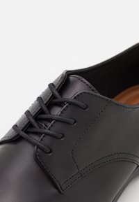 Chaussure de ville en cuir noir à lacets avec un bout arrondi et des coutures détaillées sur fond blanc.
