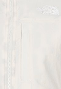 Chaqueta blanca con cremallera y un pequeño bolsillo en el pecho, con el logo de "The North Face" bordado en blanco en la parte superior izquierda. Textura suave.