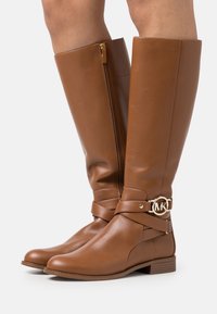 Bottes hautes en cuir marron avec fermeture éclair latérale et boucle dorée circulaire du logo MK sur une sangle, portées sur les jambes sur un fond blanc.