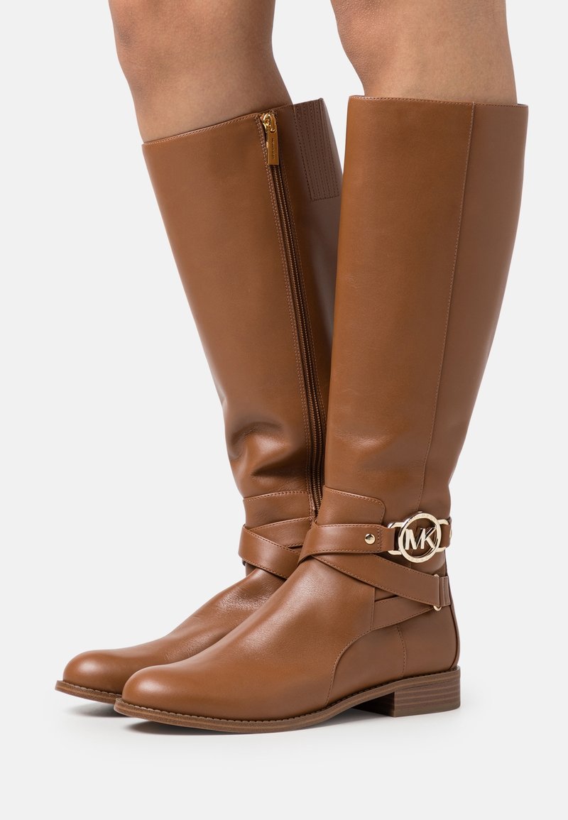 Bottes hautes en cuir marron avec fermeture éclair latérale et boucle dorée circulaire du logo MK sur une sangle, portées sur les jambes sur un fond blanc.