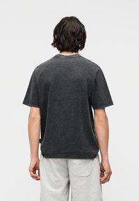 Άτομο με μεσαίου μήκους σκούρα μαλλιά, που φοράει φθαρμένο μαύρο κοντομάνικο μπλουζάκι και ανοιχτό γκρι σορτς, με την πλάτη προς την κάμερα.