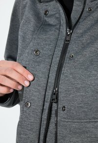 Grå zip-up jacka med texturerad tyg, snapsknappar och synlig dragkedja. En hand visas som håller jackans öppning.