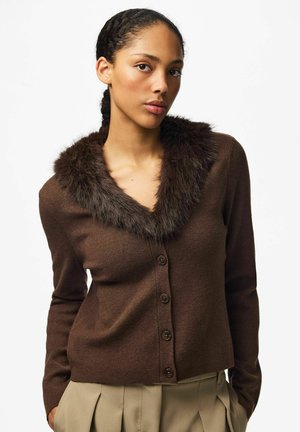 Une femme porte un cardigan marron boutonné sur le devant, avec un col en fourrure foncée et un pantalon plissé beige, sur un fond blanc uni.