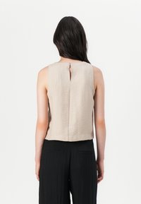 Beiges ärmelloses Oberteil mit strukturierter Oberfläche, das eine Schlüsselloch-Detail am Rücken aufweist. Kombiniert mit locker sitzenden schwarzen, hoch taillierten Hosen.