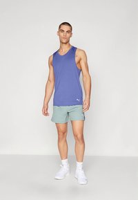 Paarse mouwloze sporttanktop, gecombineerd met lichtgroene korte broek. Voorzien van een logo, gedragen met witte sneakers en enkelsokken.