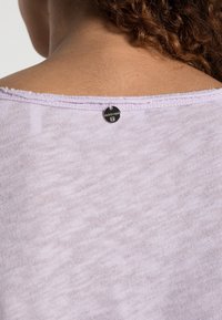 Ljust lila, mjukt tyg med en något texturerad yta. En rund, svart etikett med "rich & royal" är synlig nära halslinningen.