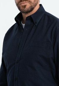 Camisa de color azul marino, de botones y con cuello, que incluye un bolsillo en el pecho, una textura sutil y un detalle de costura en contraste.