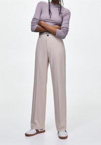 Pantalon beige pâle à jambes larges avec une taille haute et une fermeture à un seul bouton, associé à un haut ajusté à manches longues lavande et des baskets blanches.