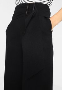 Černé široké kalhoty s vysokým pasem, vybavené páskem s obdélníkovou sponou a bočními kapsami, vyrobené z hladké texturované tkaniny.