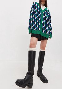 Person som bär en cardigan med geometriskt mönster, svart kjol, vita strumpor, knälda svarta stövlar och lager av halsband mot en enfärgad bakgrund.