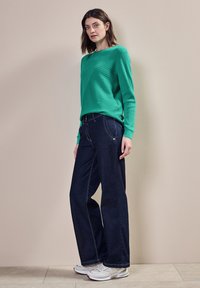 Zelený žebrovaný svetřík s dlouhým rukávem a tmavě modré široké džíny. Bílé tenisky s šedými akcenty. Jednoduchý design a volný střih.