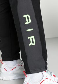 Pantaloni della tuta neri e grigi presentano una grafica verticale "AIR" in verde chiaro sul lato. Abbinati a sneakers bianche e calzini bianchi.
